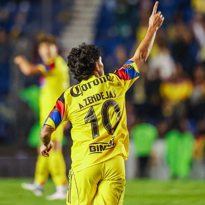 Alejandro Zendejas con una camiseta de fútbol amarilla con el texto "Corona" y "Zendejas" en la espalda, junto con el número 10. Está en un campo de hierba, levantando su brazo derecho en celebración. Otro jugador con una camiseta amarilla similar es visible en el fondo, con una multitud en las gradas.