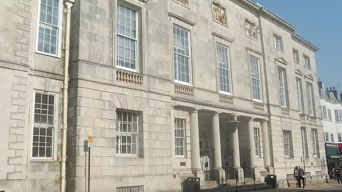 A large stone building with multiple windows and columns, identified as Lewes Court. The facade features symmetrical architecture with tall, rectangular windows and a central entrance with decorative elements above it. People are visible near the entrance, standing on the pavement.