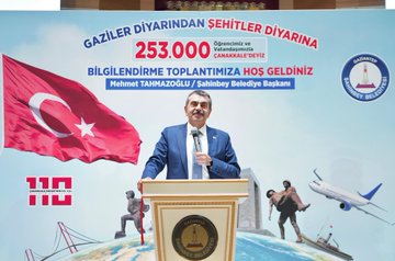 A large group of people, including students and adults, holding Turkish flags in an auditorium. Several individuals, including men in suits, stand on a stage with tables and microphones. A banner displays text and images of historical figures and an airplane. Screens and lights are visible in the background.