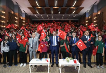 A large group of people, including students and adults, holding Turkish flags in an auditorium. Several individuals, including men in suits, stand on a stage with tables and microphones. A banner displays text and images of historical figures and an airplane. Screens and lights are visible in the background.
