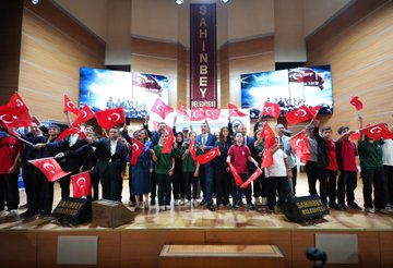 A large group of people, including students and adults, holding Turkish flags in an auditorium. Several individuals, including men in suits, stand on a stage with tables and microphones. A banner displays text and images of historical figures and an airplane. Screens and lights are visible in the background.