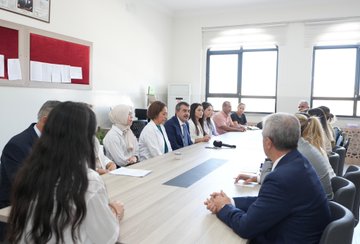 A man in a blue suit high-fiving children in yellow shirts on a school playground, with buildings in the background. Children in yellow shirts sitting at desks in a classroom, listening to a man in a blue suit and a woman. People seated around a conference table in a meeting room, including a man in a blue suit. A large group of students in yellow shirts raising their hands outside a school building with a sign reading "Emel-Galip Hasırcı Ortaokulu".