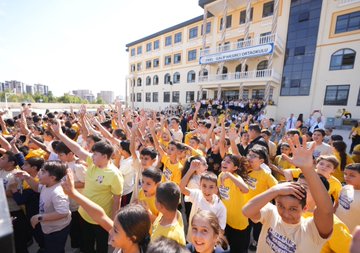 A man in a blue suit high-fiving children in yellow shirts on a school playground, with buildings in the background. Children in yellow shirts sitting at desks in a classroom, listening to a man in a blue suit and a woman. People seated around a conference table in a meeting room, including a man in a blue suit. A large group of students in yellow shirts raising their hands outside a school building with a sign reading "Emel-Galip Hasırcı Ortaokulu".