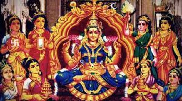 Lalitha Maha Tripurasundari seated on an ornate golden throne, adorned with blue and gold garments and jewelry. Surrounding her are multiple female figures in traditional attire, holding objects like lamps and lotuses. The background features intricate architectural elements and a golden arch.