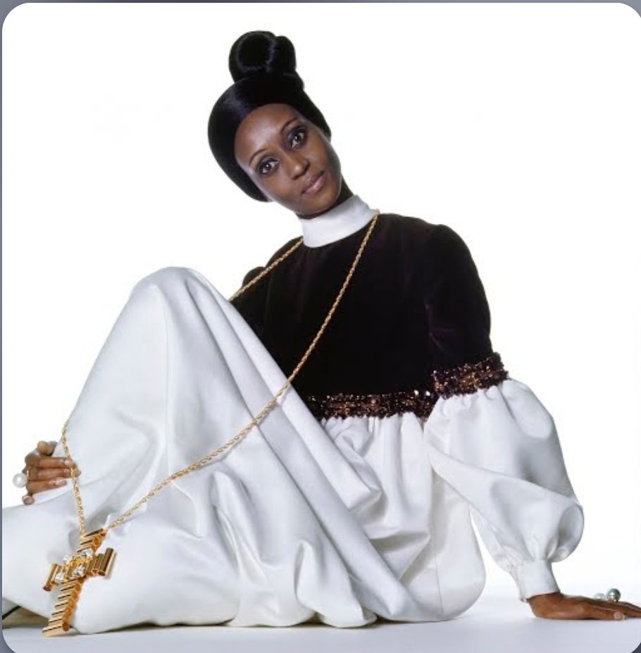From the Archive!
Model Elizabeth of Toro sitting in front of a white studio backdrop with one knee up. She is wearing an evening dress with a brown rayon velvet cropped bodice with short sleeves to the elbows that finish in full white cotton and acetate satin cuffed sleeves and