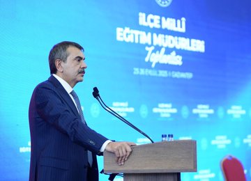A man in a dark suit stands at a wooden podium with microphones, speaking in a large conference room. Behind him, a blue backdrop displays text reading "İlçe Millî Eğitim Müdürleri Toplantısı" and "25.09.2023 Gaziantep." Another man in a gray suit holds a microphone, addressing an audience of people in formal attire seated in rows. A group of individuals in suits stands in a line in a spacious hall with red chairs and a Turkish flag visible.
