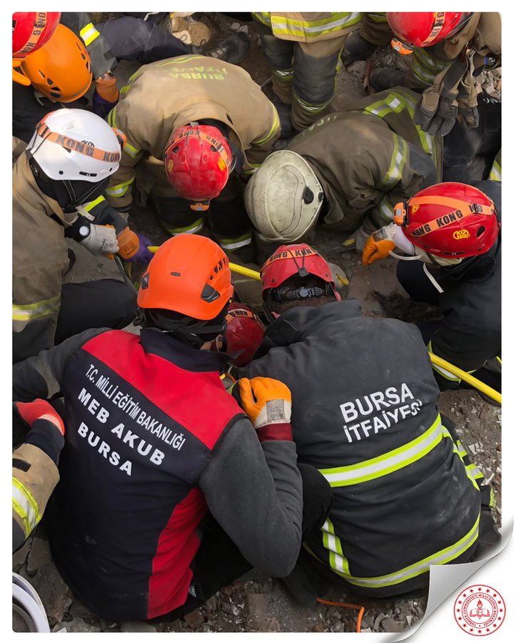 A group of firefighters wearing helmets and protective gear, including red, orange, and white helmets, gathered closely together outdoors. Some wear jackets with "BURSA İTFAİYE" and "BURSA BÜYÜKŞEHİR BELEDİYESİ İTFAİYE AKU" text. They appear to be working on a task, possibly related to an emergency response.