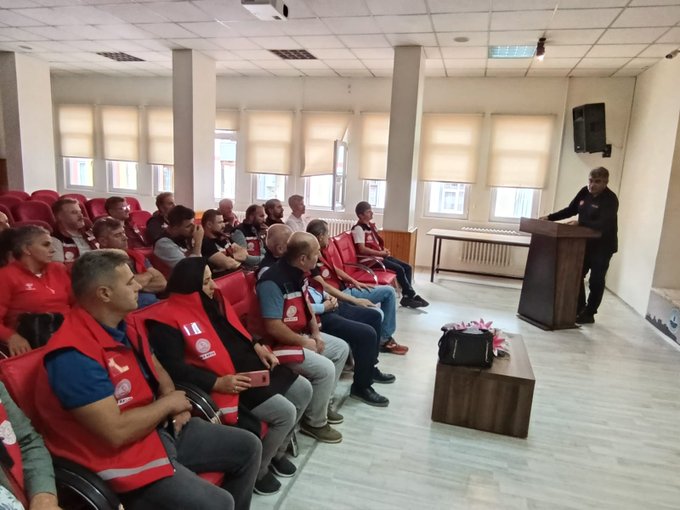 A group of people wearing red vests with white logos, sitting in rows of red chairs in a room with large windows and blinds. Some individuals stand at the front, including a person in a black suit at a podium and others in red vests. A wooden table with a bag and flowers is visible in the room. Metin Tayarer is among the group, identifiable by the context.