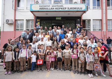 A man in a blue suit and tie giving a high-five to a young boy in a classroom, surrounded by tables with checkered tablecloths, art supplies, and students working. A woman and other children are seated at desks with notebooks and water bottles. A group of people, including men and women in formal attire, sit around a long table in a meeting room. A large group of students in uniforms and adults, including men and women, stand in front of a school building with a sign reading "Gaziantep Şehitkamil Abdullah Kepkep İlkokulu."