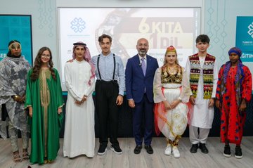 Multiple groups of people standing in a row, holding certificates and bags with a logo. Some wear traditional clothing from countries like Ethiopia, Chad, Syria, Tunisia, Albania, France, and Afghanistan. Others are dressed in formal suits or casual attire with suspenders. A backdrop displays "6 Kıta 1 Dil" and logos, with some individuals holding awards.