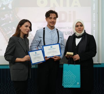 Multiple groups of people standing in a row, holding certificates and bags with a logo. Some wear traditional clothing from countries like Ethiopia, Chad, Syria, Tunisia, Albania, France, and Afghanistan. Others are dressed in formal suits or casual attire with suspenders. A backdrop displays "6 Kıta 1 Dil" and logos, with some individuals holding awards.
