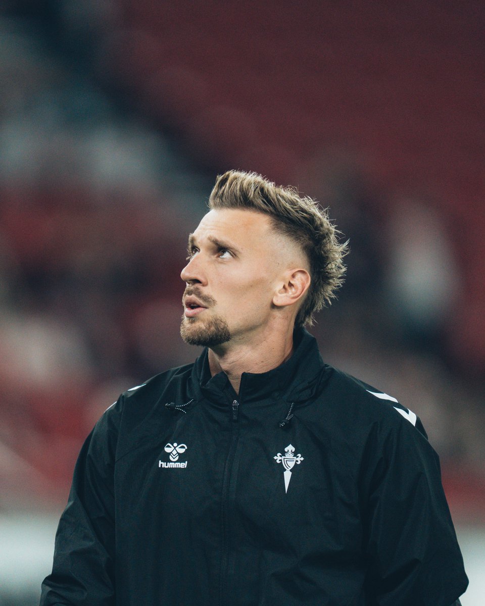 A man with blonde hair styled in a mohawk, wearing a black jacket with the Celta Vigo logo and Hummel branding. He stands in a stadium with red seating and blurred spectators in the background.