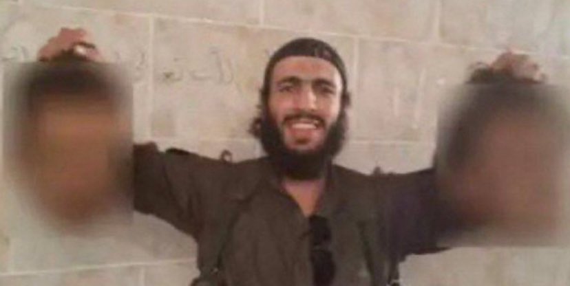 Ahmed Al Jolani holding two severed heads, standing in front of a stone wall with Arabic writing.