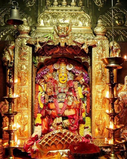 Sri Mahalakshmi Devi, adorned with a yellow face and red garments, sits in an ornate golden shrine. The statue is decorated with flowers and garlands, surrounded by intricate carvings. Oil lamps with flames illuminate the scene on either side.