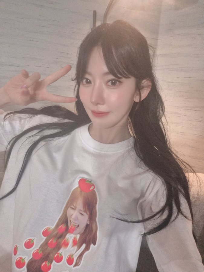Miyawaki Sakura wearing a white long-sleeve shirt with a graphic of a person with long hair and red tomatoes. She has long dark hair and is making a peace sign with her right hand. The background shows a softly lit indoor setting with a textured wall and a couch.