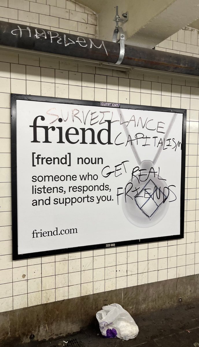 A billboard in a subway tiled with white tiles, displaying text defining "friend" as someone who listens, responds, and supports you. Graffiti reading "SURVEILLANCE FRIEND CAPITALISM" and "GET REAL FRIENDS" is scrawled over the billboard. A pipe and a bag are visible on the ground below the billboard. The website "friend.com" is printed at the bottom.
