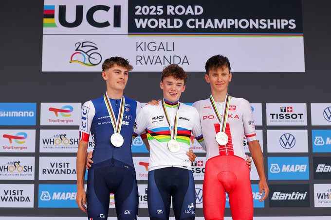 Three young male cyclists, Harry Hudson, Johan Blanc, and Jan Jackowiak, standing on a podium. Harry Hudson wears a white jersey with rainbow stripes, holding a trophy and raising one arm. Johan Blanc, in a blue jersey with "France" text, and Jan Jackowiak, in a white and red jersey with "Polska" text, stand beside him. All wear medals around their necks. A backdrop displays "UCI 2025 Road Championships Kigali Rwanda" and sponsor logos.
