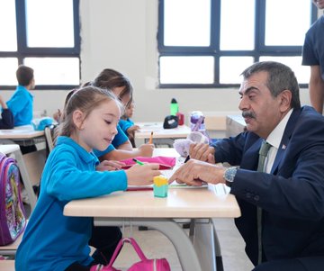 Children in blue uniforms sitting at desks in a classroom, working on activities with pencils and notebooks. A man in a suit interacts with a girl at a desk, holding a book. Other children and adults are visible, some at desks, some standing. Large windows with black frames let in natural light. A building exterior with a sign reading "Sugözü İlkokulu ve Ortaokulu" and a group of people, including men in suits and women in dresses, standing in front.