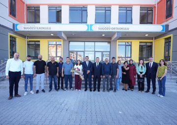 Children in blue uniforms sitting at desks in a classroom, working on activities with pencils and notebooks. A man in a suit interacts with a girl at a desk, holding a book. Other children and adults are visible, some at desks, some standing. Large windows with black frames let in natural light. A building exterior with a sign reading "Sugözü İlkokulu ve Ortaokulu" and a group of people, including men in suits and women in dresses, standing in front.
