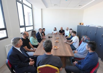 Children in blue uniforms sitting at desks in a classroom, working on activities with pencils and notebooks. A man in a suit interacts with a girl at a desk, holding a book. Other children and adults are visible, some at desks, some standing. Large windows with black frames let in natural light. A building exterior with a sign reading "Sugözü İlkokulu ve Ortaokulu" and a group of people, including men in suits and women in dresses, standing in front.