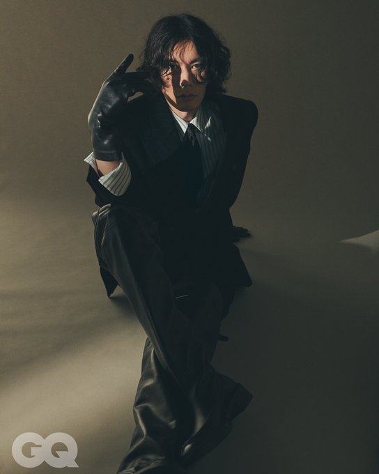 Kenshi Yonezu in a black suit and leather pants, posing in a crouched position with one hand raised, wearing black gloves. Kenshi Yonezu standing in a black suit and leather pants, with arms crossed. Both images show a neutral background with soft lighting.