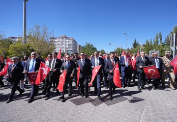 A group of people marching on a street holding Turkish flags and a white banner. Trees line the road under a clear blue sky. Men in suits carry red flags with white crescents and stars. A person in traditional orange and white attire dances in front of tents with "Besni Belediyesi" and "Merinos" text. A building displays "Ali Erdemoğlu Pen Lisesi" and posters with multiple faces.