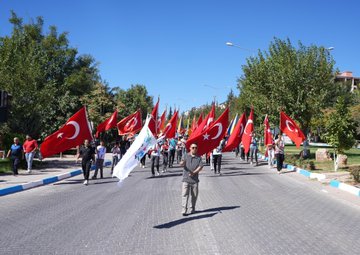 A group of people marching on a street holding Turkish flags and a white banner. Trees line the road under a clear blue sky. Men in suits carry red flags with white crescents and stars. A person in traditional orange and white attire dances in front of tents with "Besni Belediyesi" and "Merinos" text. A building displays "Ali Erdemoğlu Pen Lisesi" and posters with multiple faces.