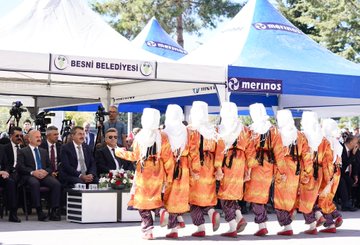 A group of people marching on a street holding Turkish flags and a white banner. Trees line the road under a clear blue sky. Men in suits carry red flags with white crescents and stars. A person in traditional orange and white attire dances in front of tents with "Besni Belediyesi" and "Merinos" text. A building displays "Ali Erdemoğlu Pen Lisesi" and posters with multiple faces.