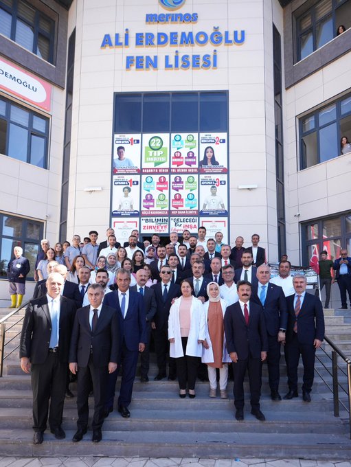 A group of people marching on a street holding Turkish flags and a white banner. Trees line the road under a clear blue sky. Men in suits carry red flags with white crescents and stars. A person in traditional orange and white attire dances in front of tents with "Besni Belediyesi" and "Merinos" text. A building displays "Ali Erdemoğlu Pen Lisesi" and posters with multiple faces.