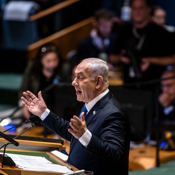 Benjamin Netanyahu se para en un podio, gesticulando con ambas manos. Viste un traje oscuro, camisa blanca y corbata azul. Los papeles y los micrófonos están en el podio. El escenario parece ser un salón de actos formal con paneles de madera y asistentes borrosos en el fondo.