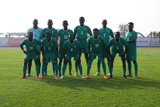 A group of soccer players wearing green jerseys with numbers 2, 3, 5, 6, 19, 20, and others, standing on a grassy field. One player in a red and white goalkeeper uniform stands to the left. The players are positioned in two rows, some standing and some kneeling.