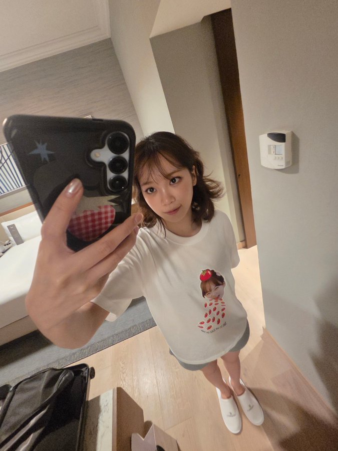 Kim Chaewon taking a mirror selfie in a hotel room. She wears a white T-shirt with a red and white character design and white shoes. Her phone, with a star sticker, is visible as she holds it for the selfie. A suitcase and bed are in the background.