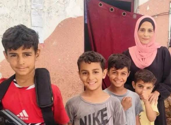 Four boys and a woman standing in front of a wall and a red curtain. The boys wear casual clothing, including a red shirt with a black strap, a gray Nike shirt, and light-colored shirts. The woman wears a pink headscarf and dark clothing.