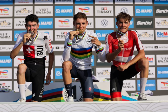 Lorenzo Finn in a white jersey with rainbow stripes, gold medal around neck, standing on top podium step, arms raised. Jan Huber in black and white jersey with Swiss flag, silver medal, on left podium step. Marco Schrettl in red and white jersey with Austrian flag, bronze medal, on right podium step. Background shows UCI logos, sponsors like Tissot and Volkswagen, and text reading "UCI 2025 Road Championships Kigali Rwanda".
