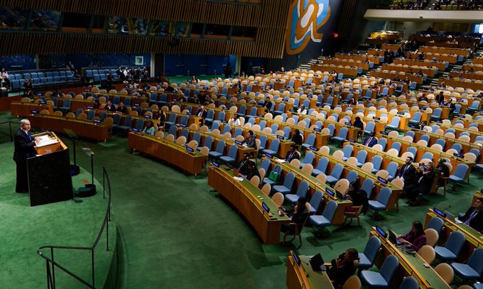 A large auditorium with rows of blue and beige chairs, mostly empty. A man stands at a podium on a green carpeted floor, speaking to a sparse audience. Wooden desks and seats are arranged in a semi-circular pattern, with a large emblem on the wall.