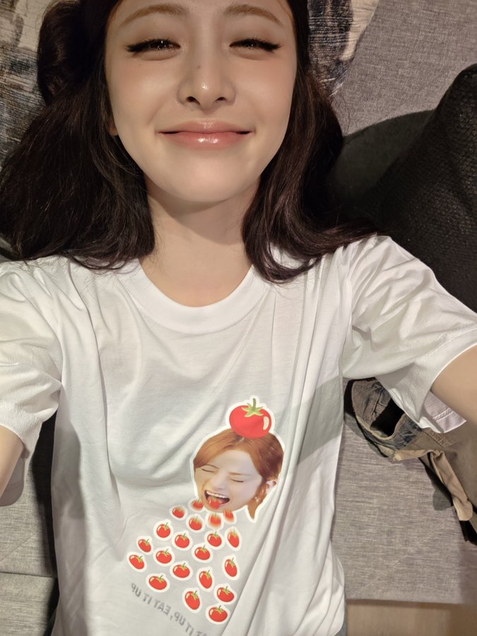 Hwang Hyunjin wearing a white T-shirt with a graphic of a person with a tomato on their head and red tomatoes falling. The person has long hair and is depicted with an open mouth. Hwang Hyunjin has shoulder-length dark hair and is lying on a gray couch, smiling at the camera.