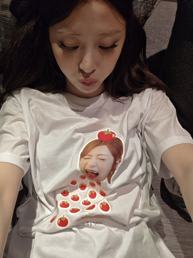 Hwang Hyunjin wearing a white T-shirt with a graphic of a person with a tomato on their head and red tomatoes falling. The person has long hair and is depicted with an open mouth. Hwang Hyunjin has shoulder-length dark hair and is lying on a gray couch, smiling at the camera.