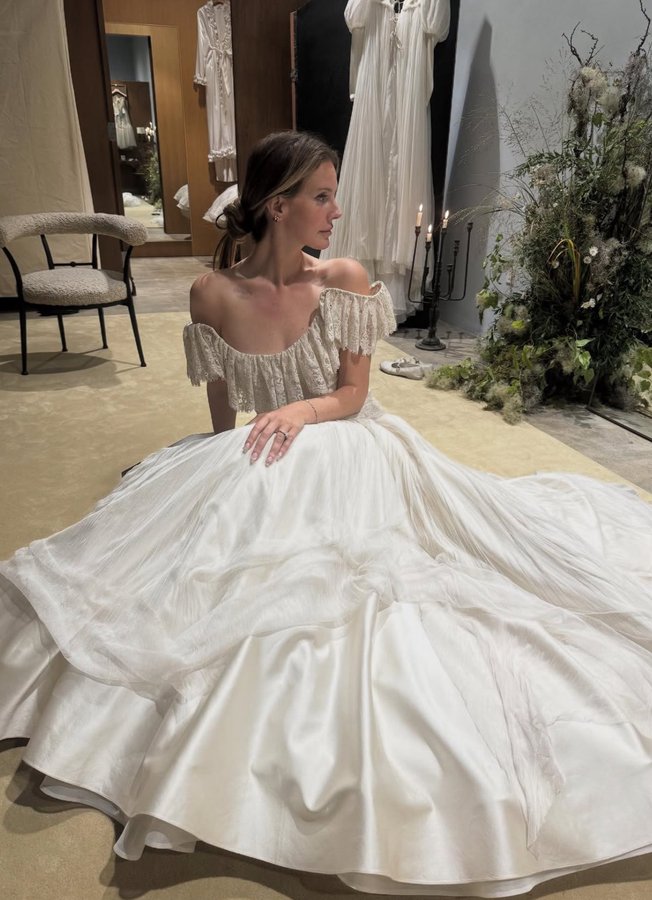 Lana Del Rey wearing a voluminous white wedding dress with off-shoulder sleeves, sitting on the floor in an elegant room with floral arrangements and dresses on racks. Jeremy in a dark suit and vest, sitting beside Lana Del Rey on a bench near an airboat at night in a black-and-white photo.