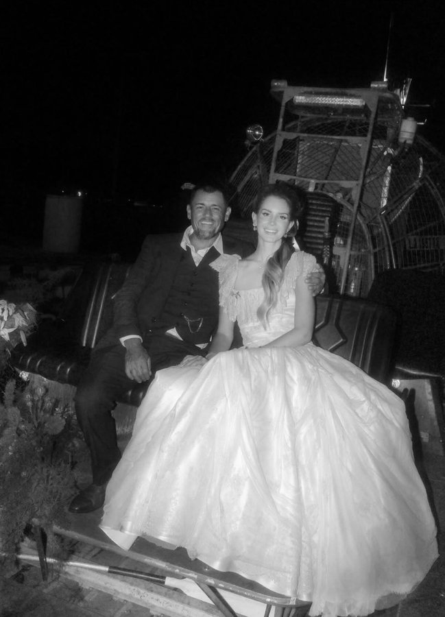 Lana Del Rey wearing a voluminous white wedding dress with off-shoulder sleeves, sitting on the floor in an elegant room with floral arrangements and dresses on racks. Jeremy in a dark suit and vest, sitting beside Lana Del Rey on a bench near an airboat at night in a black-and-white photo.