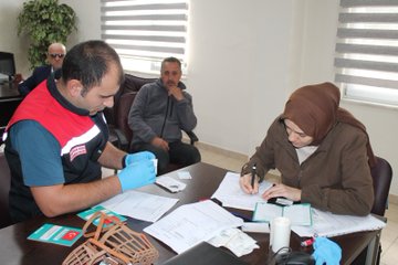 First image shows outdoor scene with people in red vests handling a dog near a table with supplies and Turkish flag banner. Second image depicts indoor office setting with man in vest wearing gloves assisting woman in headscarf at desk with documents and Turkish flag basket. Third image features men in vests examining a dog outdoors near fence and trees. Fourth image illustrates woman and another in headscarf with gloves vaccinating small dog on table in room with posters of animals.