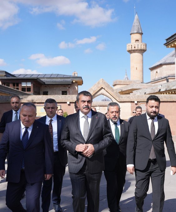 First image shows a group of men in dark suits and ties standing outdoors in front of a tall beige minaret and historic stone buildings with arched doorways under a clear blue sky with scattered clouds. Second image depicts a stone fountain with a carved lion head spouting water into a basin, Arabic inscriptions on the surrounding arched stone wall, and a man in a dark suit and glasses bending down to drink from the stream while others in casual dark clothing observe nearby. Third image features several men in suits gathered around a glass display case containing an open ancient Quran manuscript with ornate script, set in an indoor room with white walls, golden Arabic plaques, and green carpet flooring. Fourth image displays a group of eight people including men in suits and two women in black dresses and scarves standing in a line outdoors in front of a large arched stone entranceway with a Turkish flag emblem above, on a paved area with greenery in the background.
