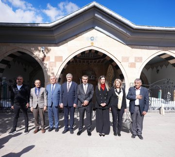 First image shows a group of men in dark suits and ties standing outdoors in front of a tall beige minaret and historic stone buildings with arched doorways under a clear blue sky with scattered clouds. Second image depicts a stone fountain with a carved lion head spouting water into a basin, Arabic inscriptions on the surrounding arched stone wall, and a man in a dark suit and glasses bending down to drink from the stream while others in casual dark clothing observe nearby. Third image features several men in suits gathered around a glass display case containing an open ancient Quran manuscript with ornate script, set in an indoor room with white walls, golden Arabic plaques, and green carpet flooring. Fourth image displays a group of eight people including men in suits and two women in black dresses and scarves standing in a line outdoors in front of a large arched stone entranceway with a Turkish flag emblem above, on a paved area with greenery in the background.