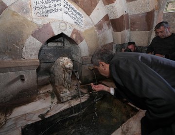 First image shows a group of men in dark suits and ties standing outdoors in front of a tall beige minaret and historic stone buildings with arched doorways under a clear blue sky with scattered clouds. Second image depicts a stone fountain with a carved lion head spouting water into a basin, Arabic inscriptions on the surrounding arched stone wall, and a man in a dark suit and glasses bending down to drink from the stream while others in casual dark clothing observe nearby. Third image features several men in suits gathered around a glass display case containing an open ancient Quran manuscript with ornate script, set in an indoor room with white walls, golden Arabic plaques, and green carpet flooring. Fourth image displays a group of eight people including men in suits and two women in black dresses and scarves standing in a line outdoors in front of a large arched stone entranceway with a Turkish flag emblem above, on a paved area with greenery in the background.