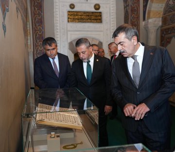 First image shows a group of men in dark suits and ties standing outdoors in front of a tall beige minaret and historic stone buildings with arched doorways under a clear blue sky with scattered clouds. Second image depicts a stone fountain with a carved lion head spouting water into a basin, Arabic inscriptions on the surrounding arched stone wall, and a man in a dark suit and glasses bending down to drink from the stream while others in casual dark clothing observe nearby. Third image features several men in suits gathered around a glass display case containing an open ancient Quran manuscript with ornate script, set in an indoor room with white walls, golden Arabic plaques, and green carpet flooring. Fourth image displays a group of eight people including men in suits and two women in black dresses and scarves standing in a line outdoors in front of a large arched stone entranceway with a Turkish flag emblem above, on a paved area with greenery in the background.