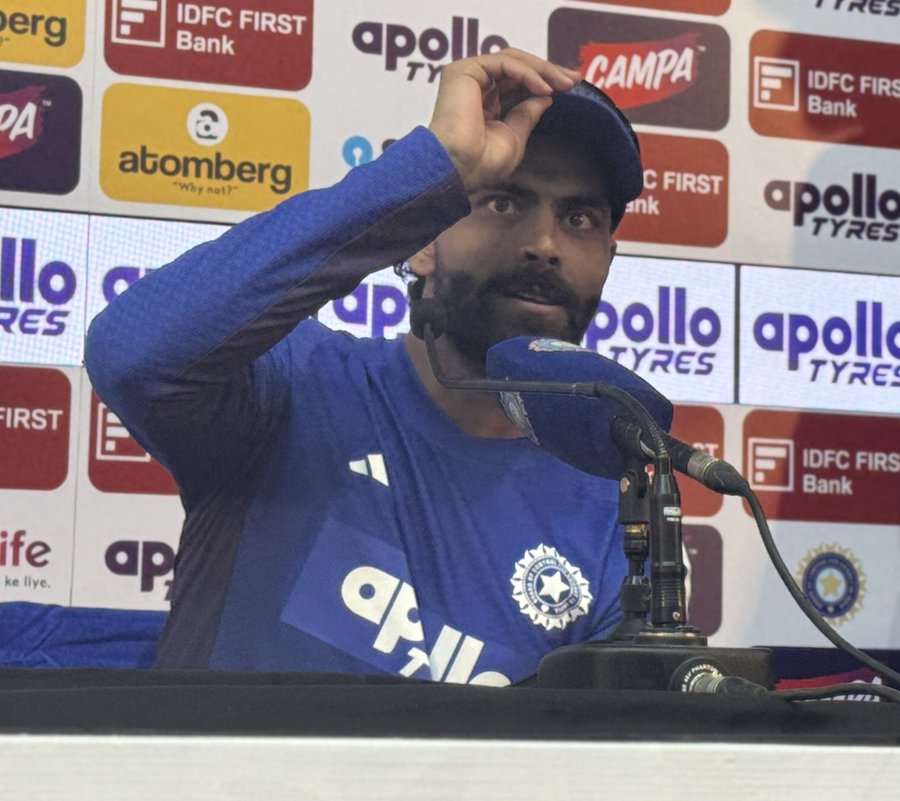A man wearing a blue cricket jersey with Apollo sponsor logo and BCCI emblem stands at a press conference podium surrounded by sponsor backdrops including IDFC First Bank, Apollo Tyres, and Atomberg. He holds a cap on his head with his right hand while speaking into a microphone held in his left hand. The setting features multiple sponsor banners in yellow, red, and blue colors.