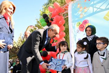 First image shows a diverse group of people including men and women in formal attire and headscarves sitting on blue chairs under a canopy, engaged in prayer with hands raised, surrounded by autumn trees and umbrellas in an outdoor setting. Second image depicts a large crowd of attendees waving Turkish and Palestinian flags on a grassy field with tents and high-rise buildings in the background under a clear sky. Third image captures a man in a suit handing out striped gift bags to smiling children dressed in colorful outfits near balloons and trees. Fourth image features a group of suited officials and young girls in dresses holding medals on stage with a banner reading Malatya Müftülüğü Bed-i Besmele, decorated with green and red balloons.