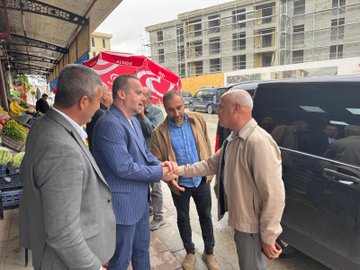 First image shows five middle-aged men in suits and casual attire sitting around a low wooden table on a street outside a brick building with large green watermelons and pumpkins in crates nearby, some holding tea glasses and chatting. Second image depicts six men in business suits seated at a similar low table in front of a market shop window displaying fruits and vegetables like bananas and greens, with a red and white umbrella visible and tea glasses on the table. Third image captures four men in suits shaking hands on a street near a market stall with flowers and produce crates, beside a black van and under a canopy with a red umbrella.