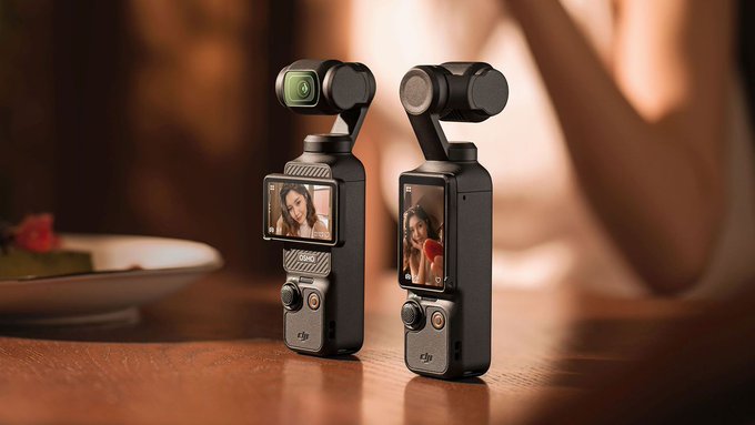 Two black DJI Osmo Pocket devices placed on a wooden table. The left device shows a front view with a small circular lens and a screen displaying a woman with long dark hair in a close-up portrait. The right device is in side view with a rectangular screen showing the same woman in a similar portrait. A blurred background includes a plate with food and a persons arm.