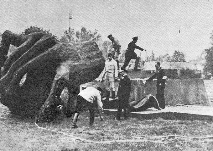 Black-and-white photograph showing several people in 1940s attire including men in suits hats and uniforms and women in dresses gathered around a large fallen concrete structure that appears to be a toppled monument or statue base with irregular stone shapes resembling limbs or abstract forms partially buried in grass under trees with a flagpole and another pole in the background the scene suggests destruction or removal of a public artwork in an outdoor urban park setting.