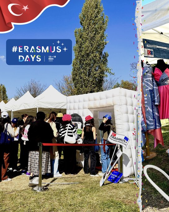 Outdoor event setup in a park with green grass and trees under blue sky. White tents and inflatable structures with Erasmus+ Days banners in blue and yellow. Turkish flag waving on left. People in casual clothes including hats and striped shirts gathered around barriers and tables with promotional items like pillows and documents. Colorful displays with EU flags and rainbow elements on tables. Photo booth area with props like signs and flowers in pink and purple. Additional tent with red tablecloth and blue materials.